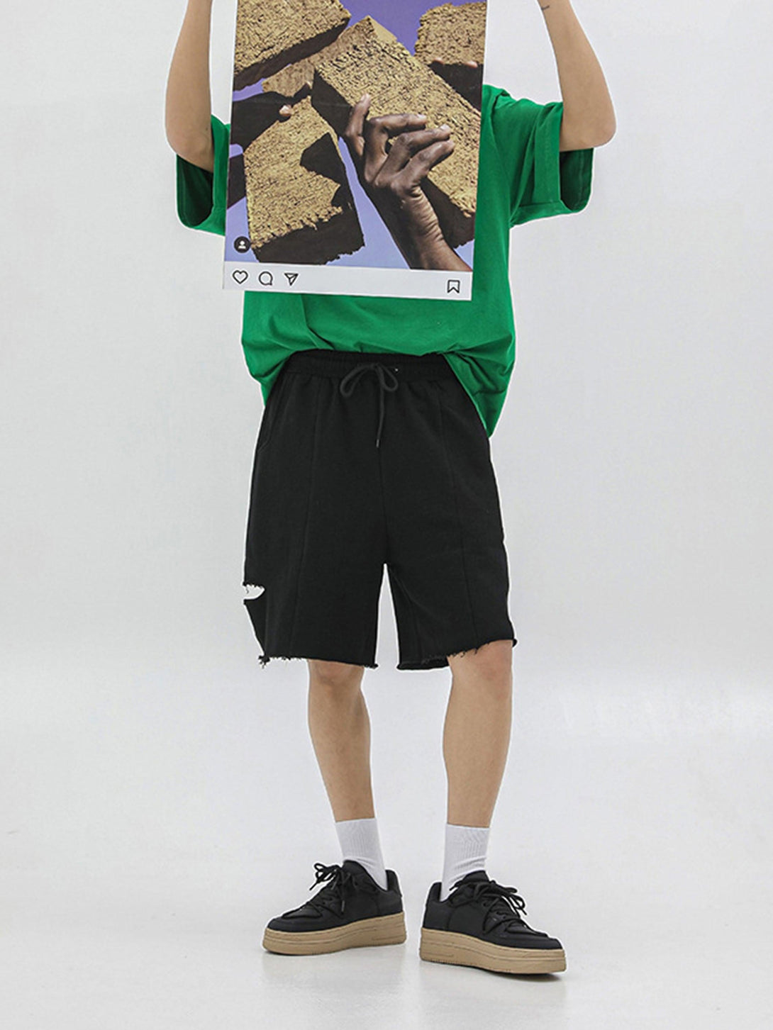 Model in green shirt and black shorts holding artwork, wearing black TALISHKO sneakers.