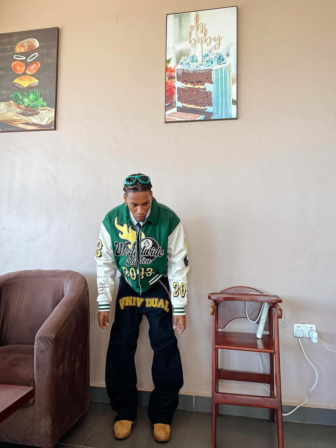 Model in TALISHKO varsity jacket and jeans, styled with casual boots in a cozy indoor setting.