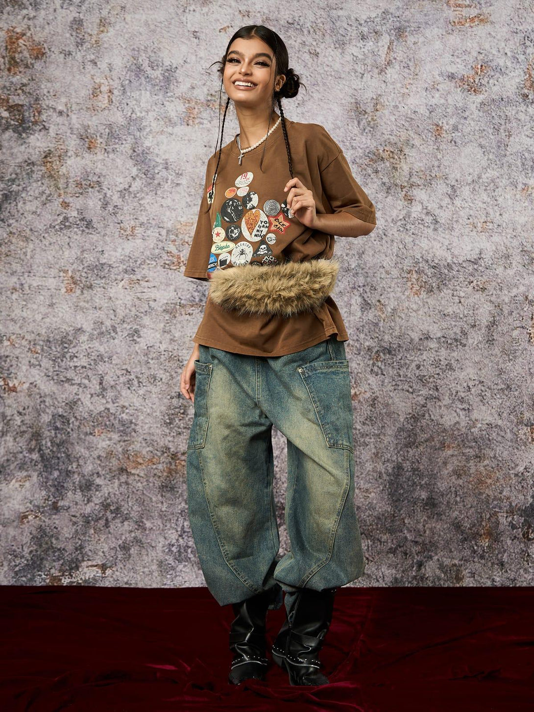 Model in a brown TALISHKO T-shirt with badge print, paired with denim and boots for a trendy ensemble.