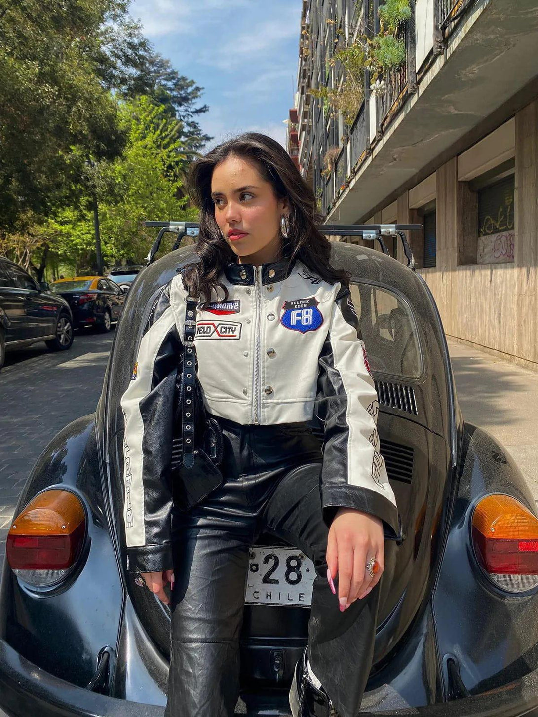 Model in TALISHKO's PU leather racing jacket with badges, seated on a classic car in urban setting.