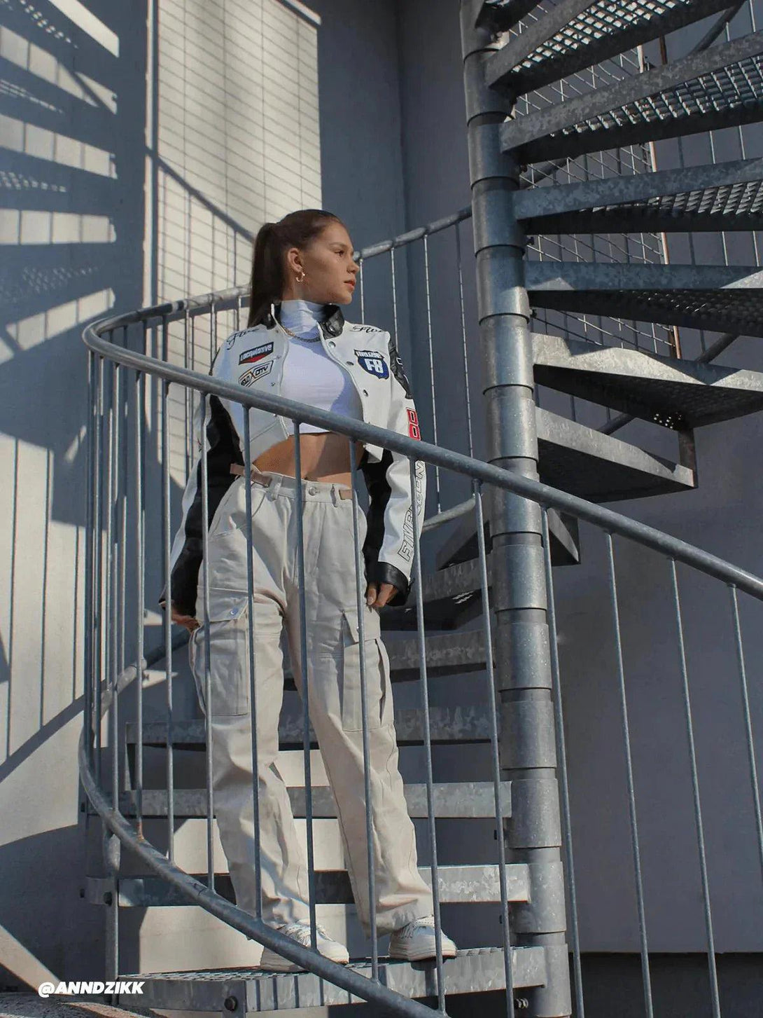 Model in TALISHKO's badge-adorned racing jacket on a spiral staircase, exuding urban style.