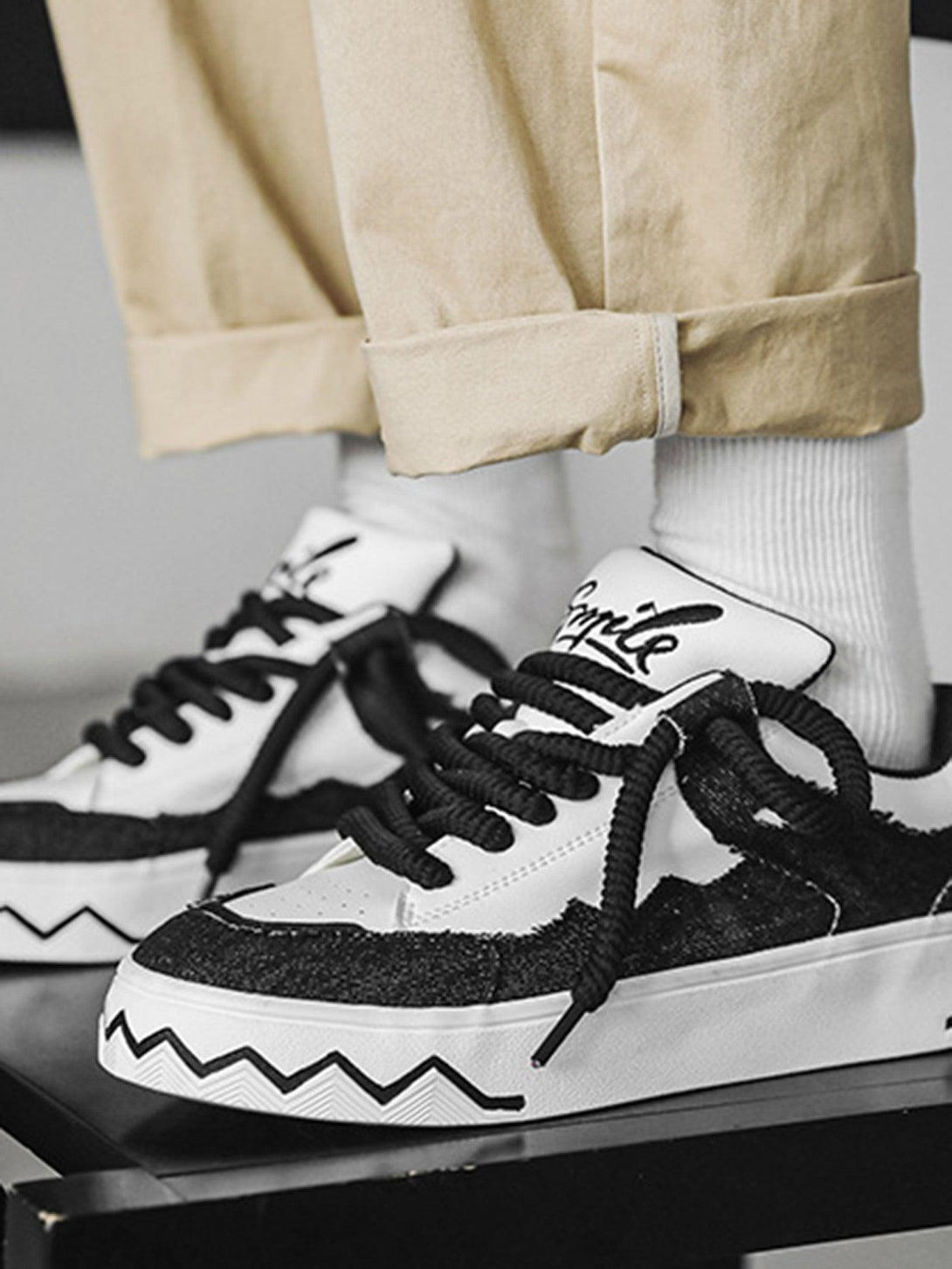 Model wearing TALISHKO sneakers with bold zigzag soles and black-white contrast, paired with beige pants.