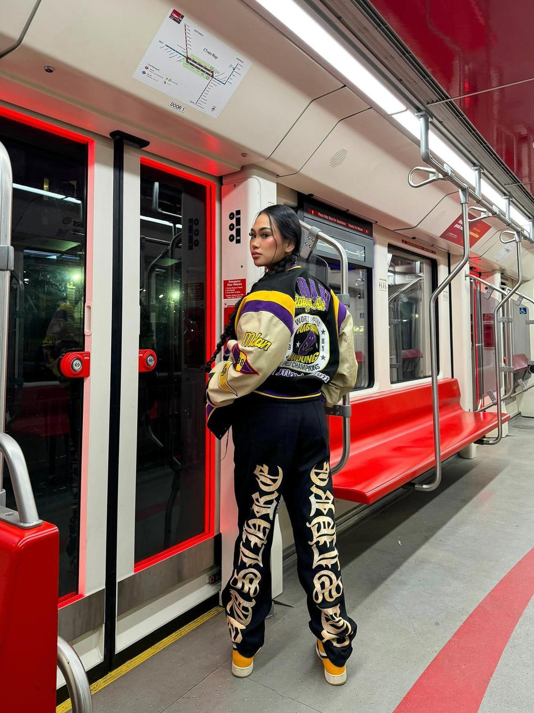 Model in TALISHKO jeans with bold lettering, styled with a varsity jacket in a subway setting.