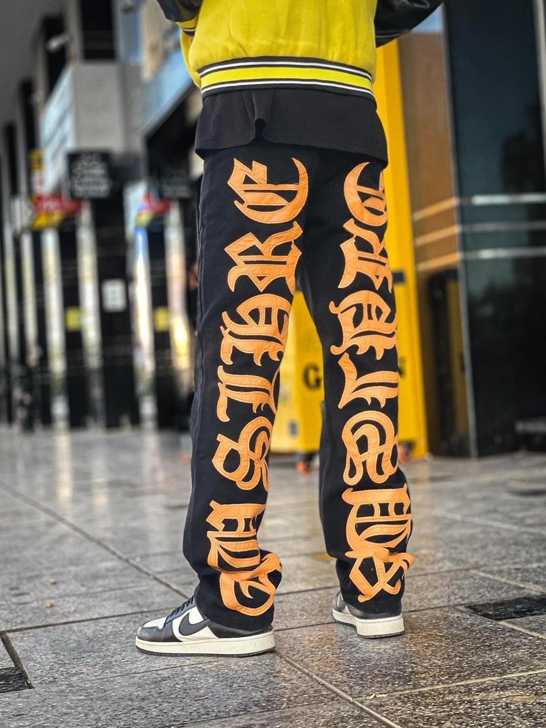 Back view of TALISHKO jeans with bold orange embroidery, styled with sneakers for a streetwear vibe.