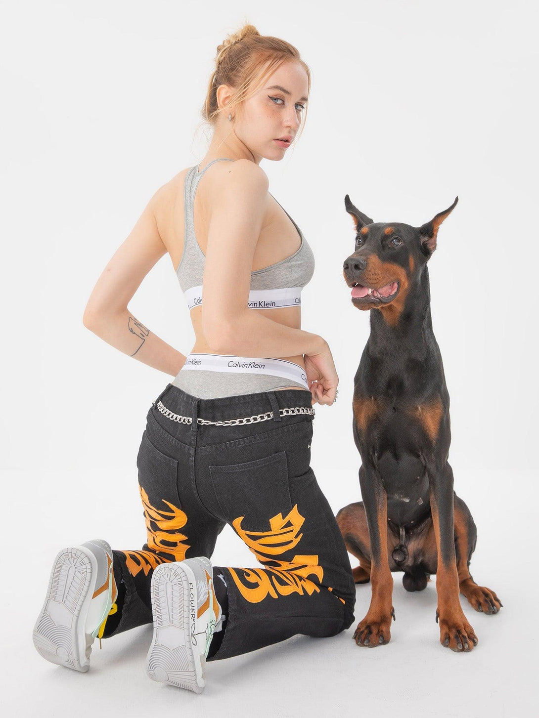 Model kneeling in TALISHKO jeans with bold orange embroidery, paired with a sporty top and sneakers.