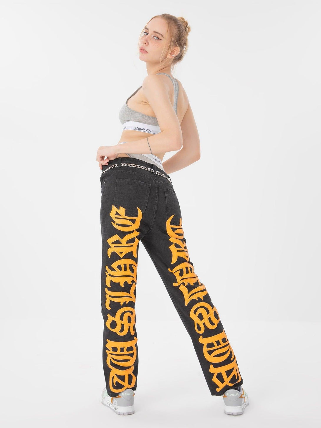 Model wearing TALISHKO black jeans with bold orange embroidered text, styled with a sporty top.