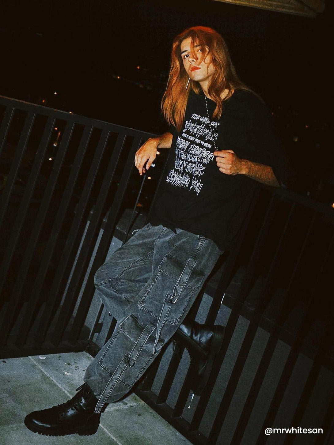 Model leans against railing in TALISHKO distressed tee with chain detail, exuding urban night style.