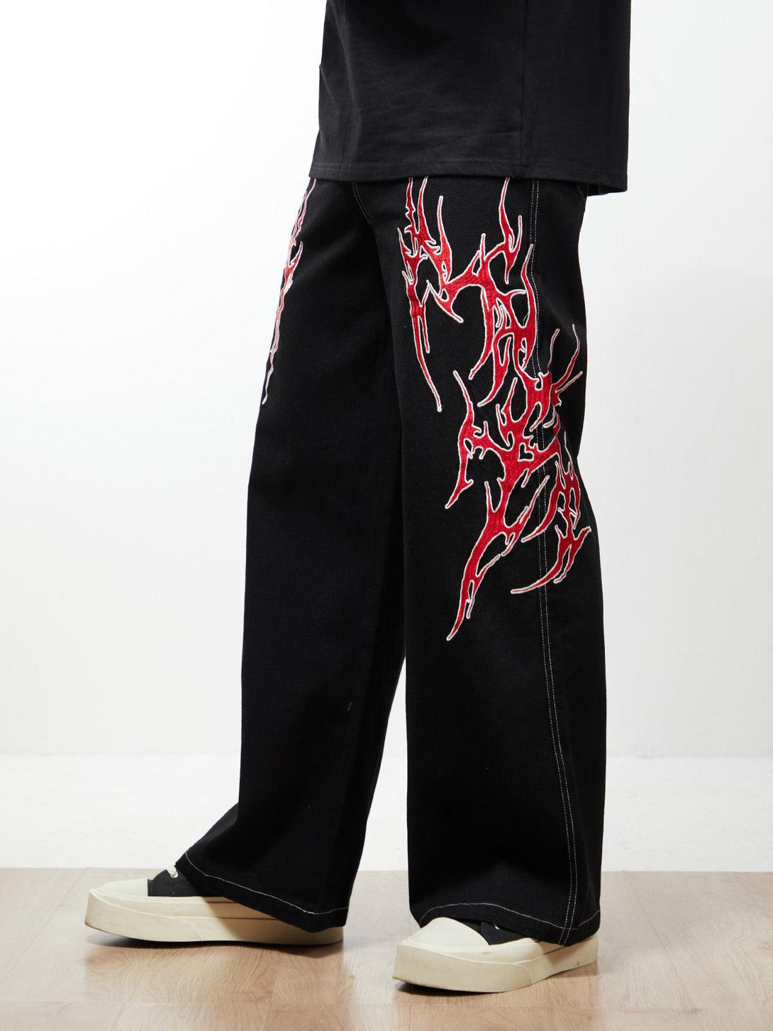 Side perspective of TALISHKO's black baggy jeans with striking red flame embroidery, paired with white sneakers.