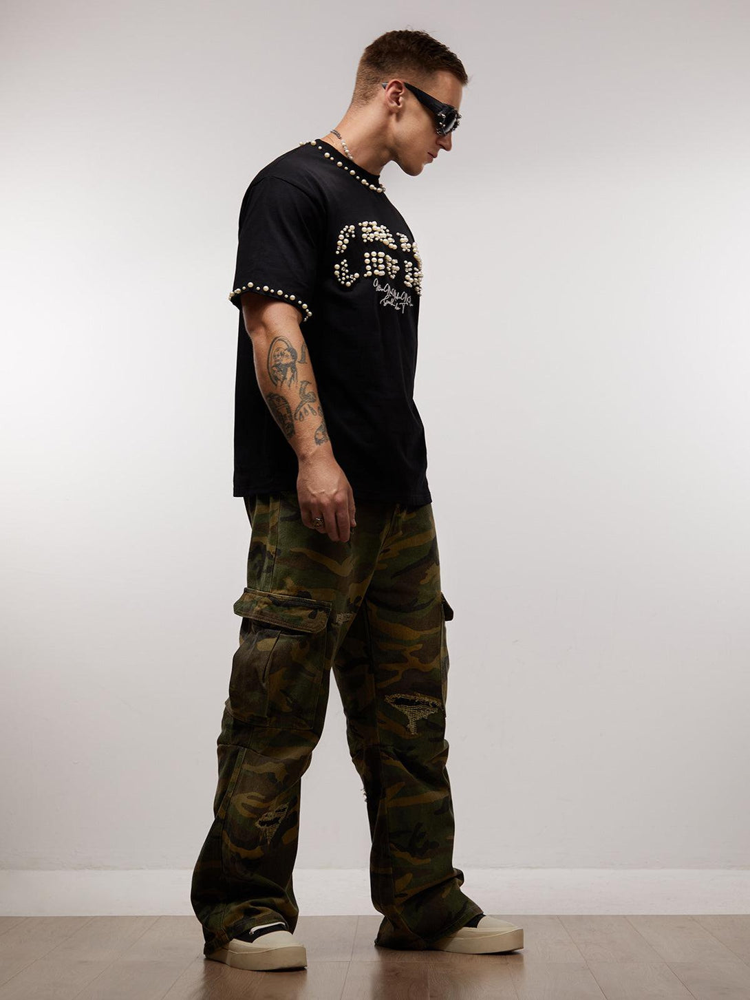 Model in pearl-embellished black T-shirt by TALISHKO and camo cargo pants, showcasing trendy streetwear.