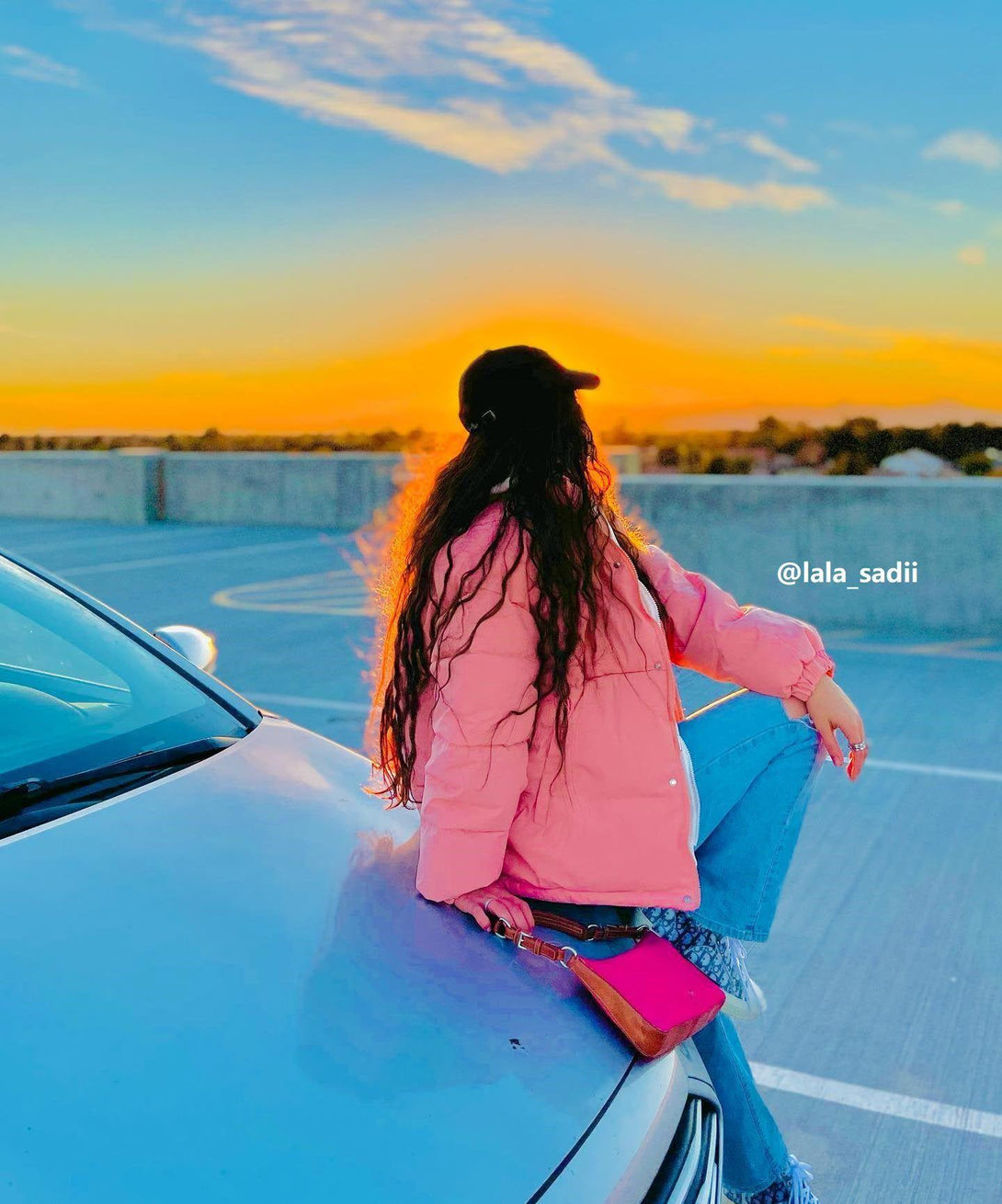 Model sitting on car hood wearing TALISHKO pink winter coat, styled with jeans and a cap.