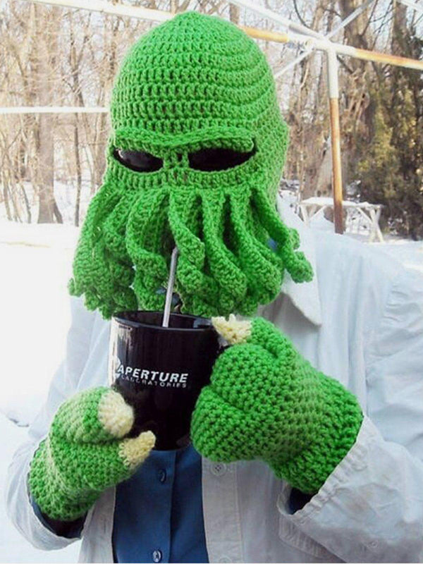 Person wearing a green knit octopus hat and gloves by TALISHKO, holding a black mug outdoors.