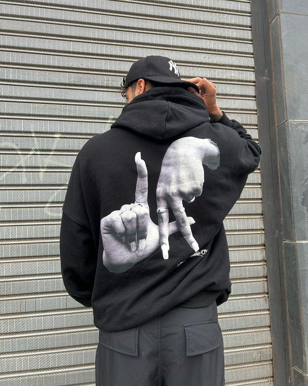 Man wearing TALISHKO Gesture Print Hoodie with striking hand graphic against a metal backdrop.