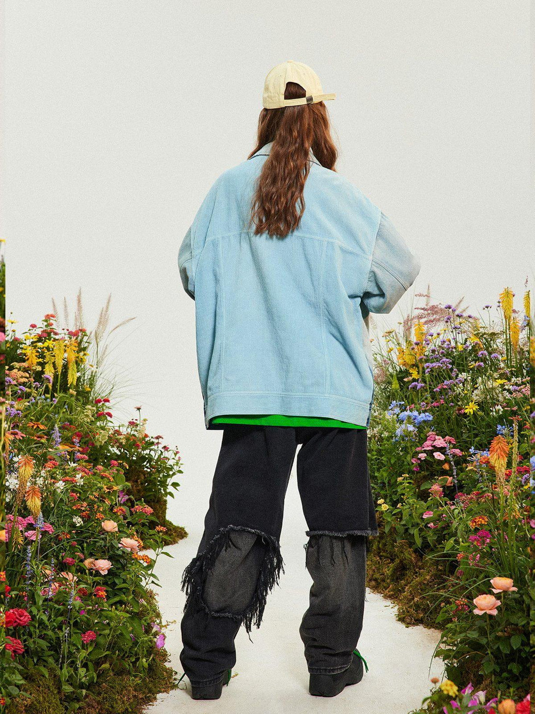Model in a TALISHKO gradient denim jacket, standing amidst vibrant garden flowers, showcasing a casual look.