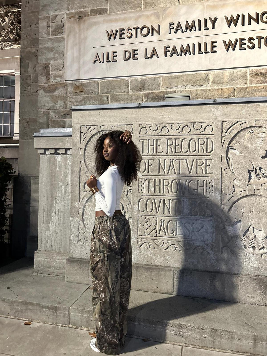 Model wearing TALISHKO camouflage pants with a white top, set against a stone backdrop.
