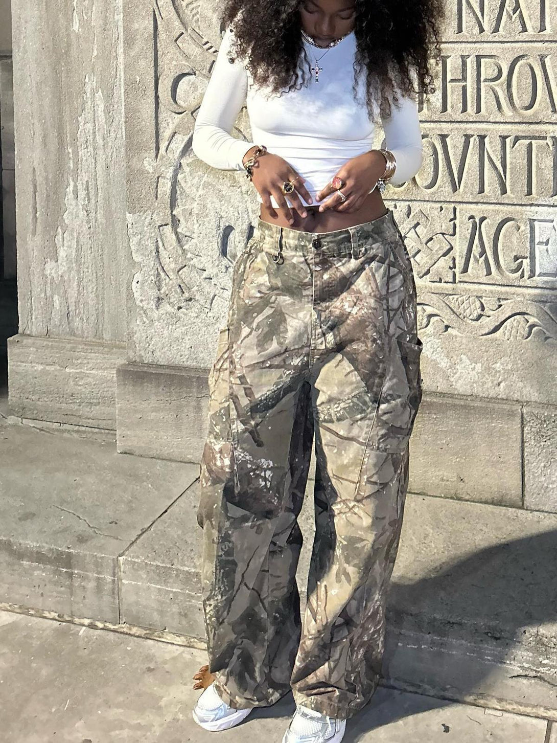 Model wearing TALISHKO camouflage pants with a white top, standing against a carved stone wall.