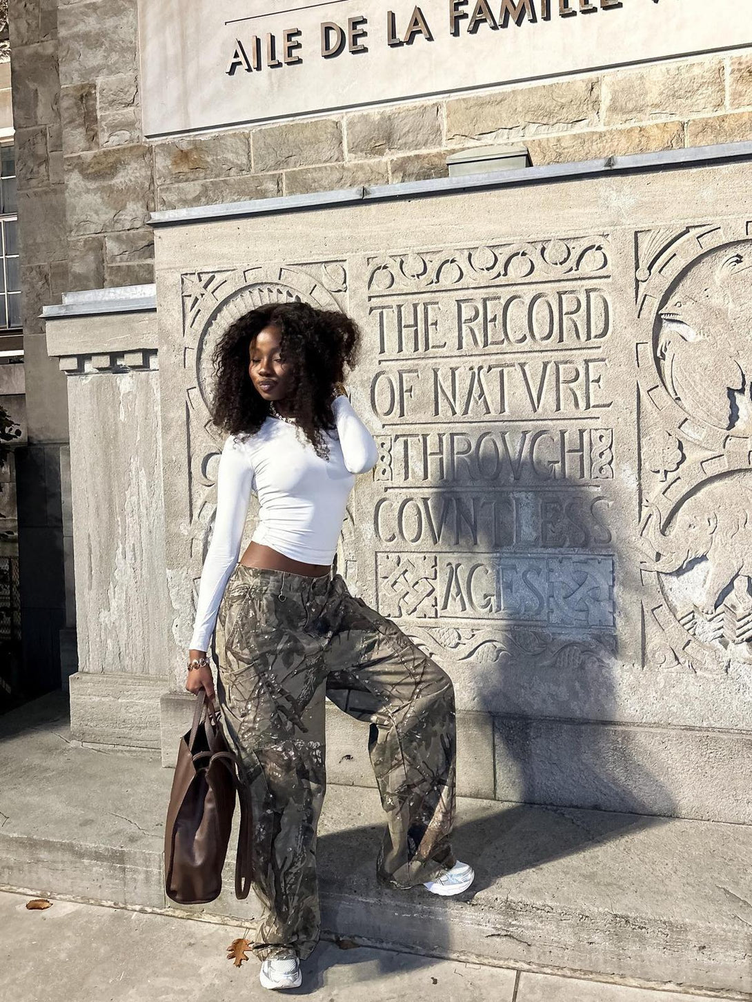 Model in TALISHKO camouflage pants and white top, posing against carved stone backdrop.