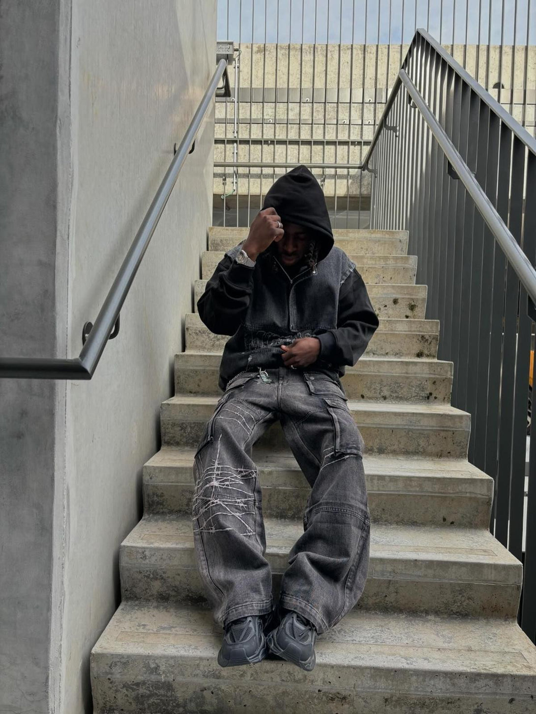 Model sitting on stairs in TALISHKO hip-hop jeans with barbed wire embroidery detailing.