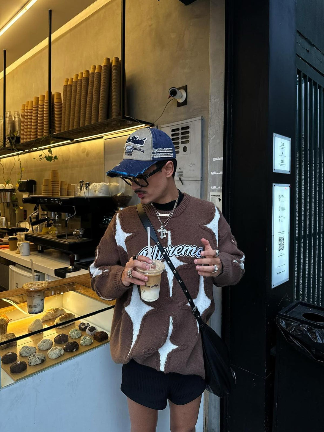 Model in TALISHKO's brown star-pattern sweater, sipping iced coffee outside a cafe.