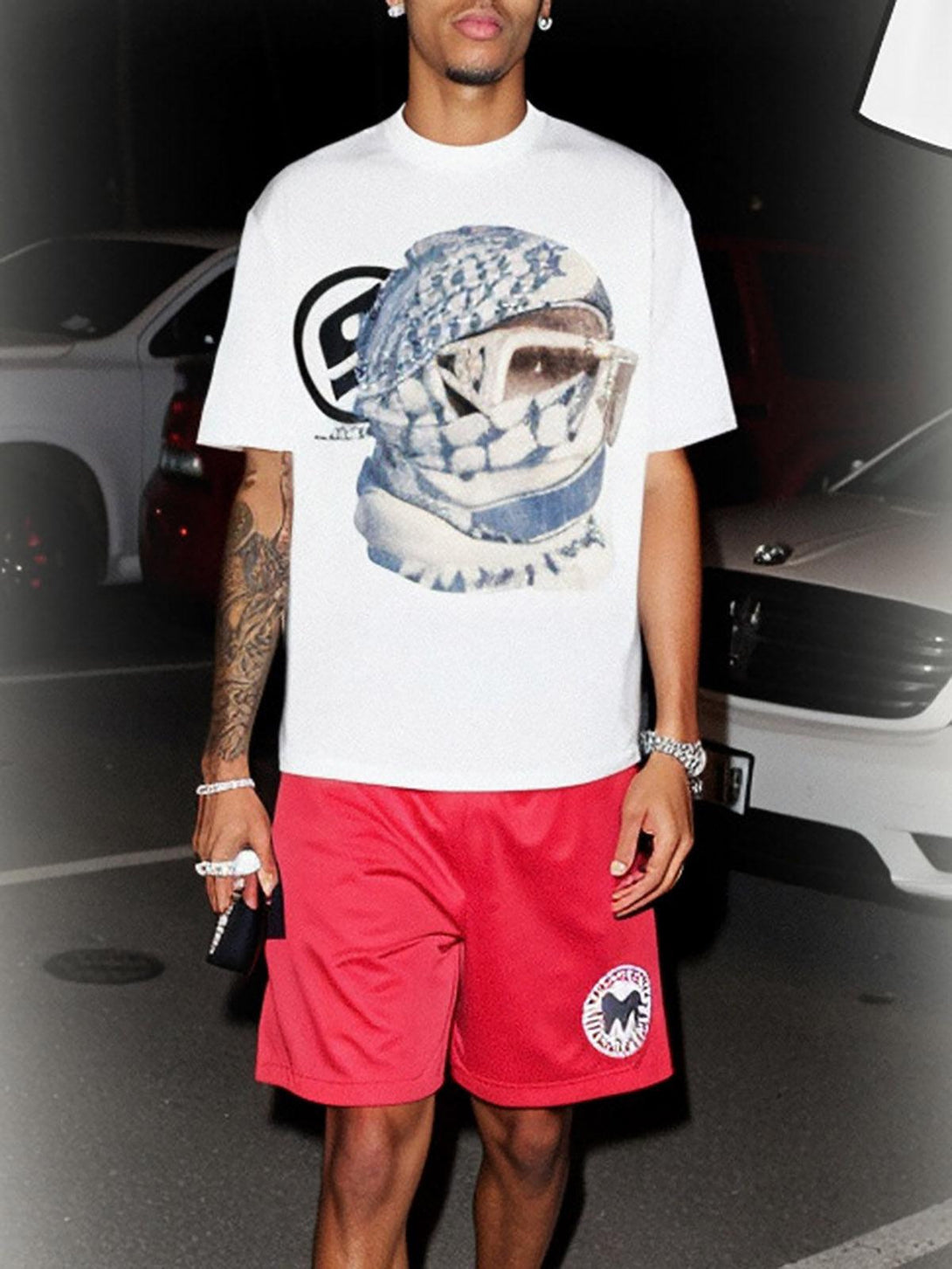Model wearing TALISHKO's white graphic T-shirt with a masked portrait, paired with red shorts.