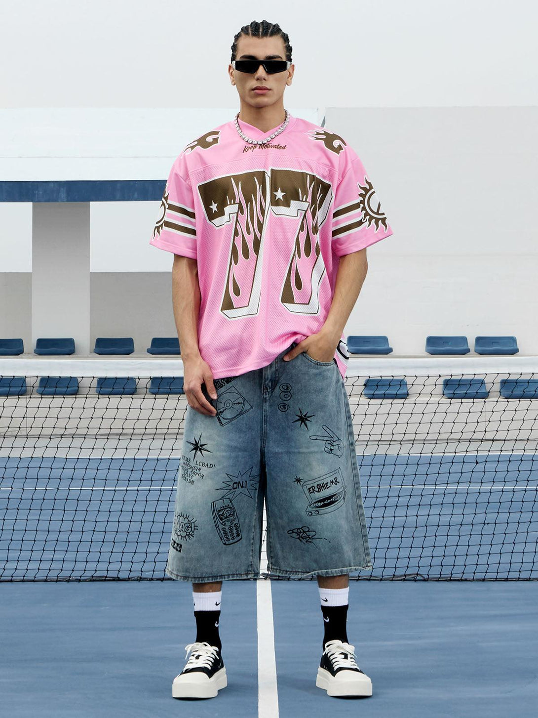 Model in a pink flame print jersey T-shirt by TALISHKO, paired with denim shorts on a tennis court.