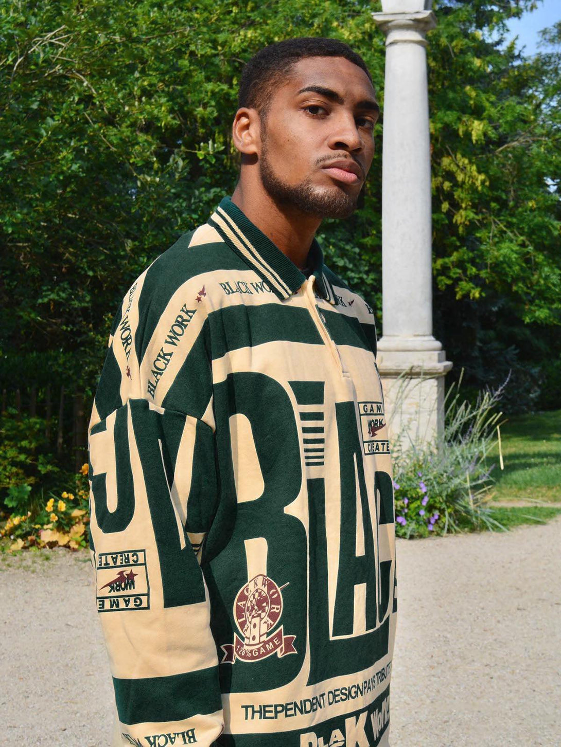 Model wearing a TALISHKO retro typography polo tee, set against a lush garden backdrop.