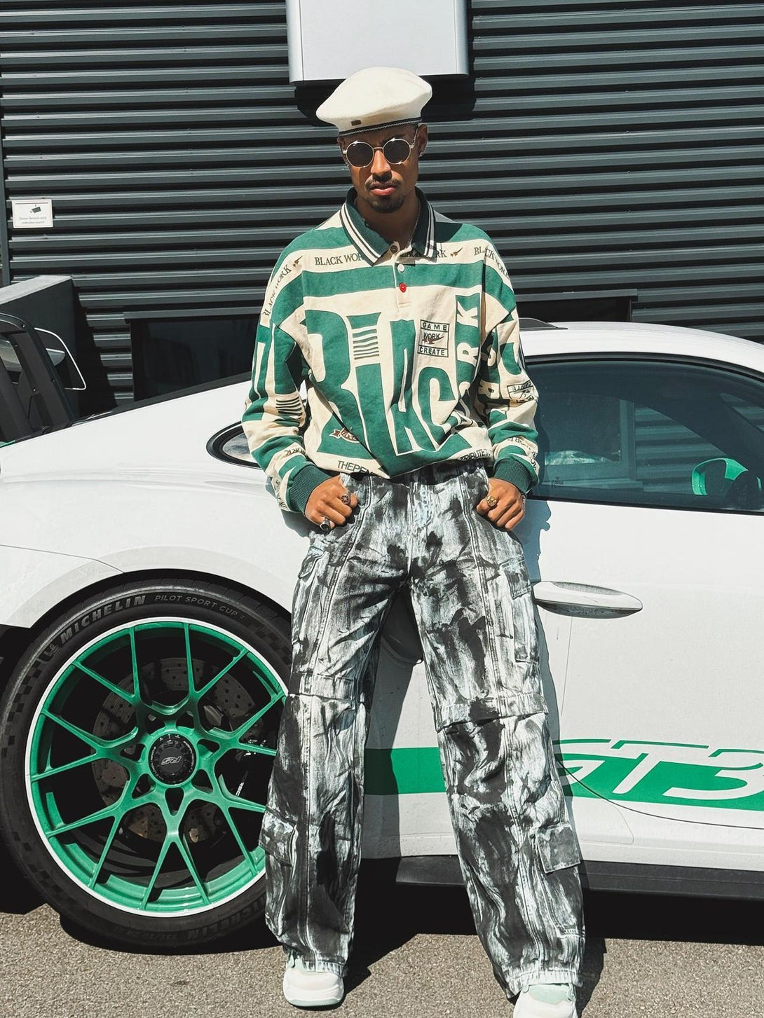 Model in a TALISHKO retro typography polo tee and artistic pants, standing by a stylish car.