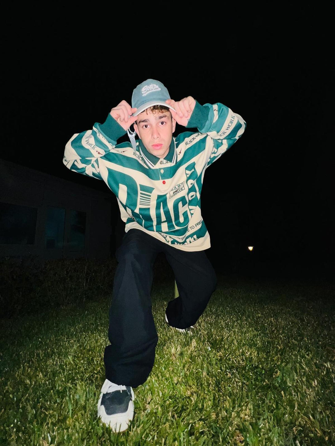 Model wearing a TALISHKO retro typography polo tee, adjusting a cap while crouching on grass.