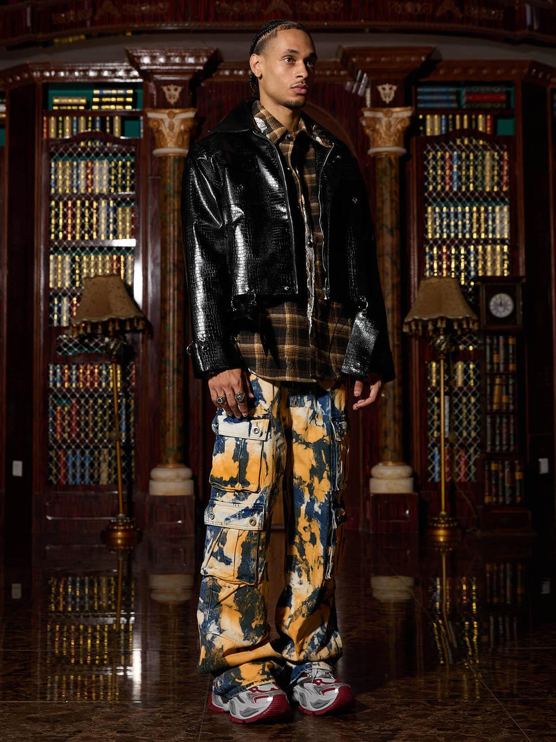 Model in TALISHKO's leather jacket paired with plaid shirt and bold cargo pants, set in a library.