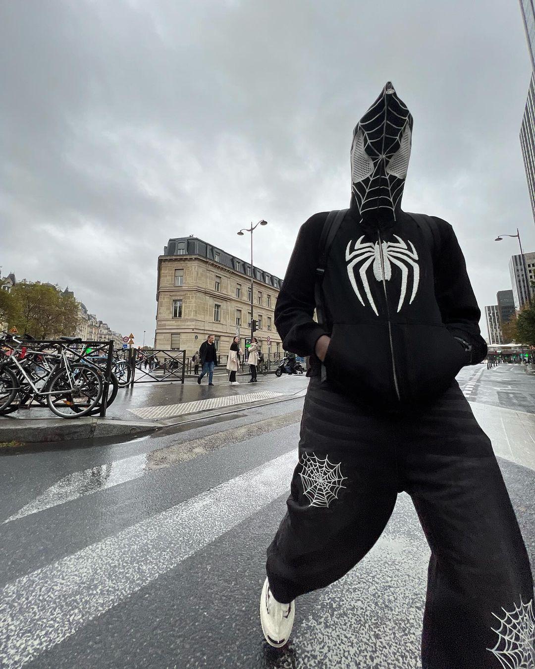 Person wearing a hooded TALISHKO outfit with spider web embroidery, standing on a rainy city street.