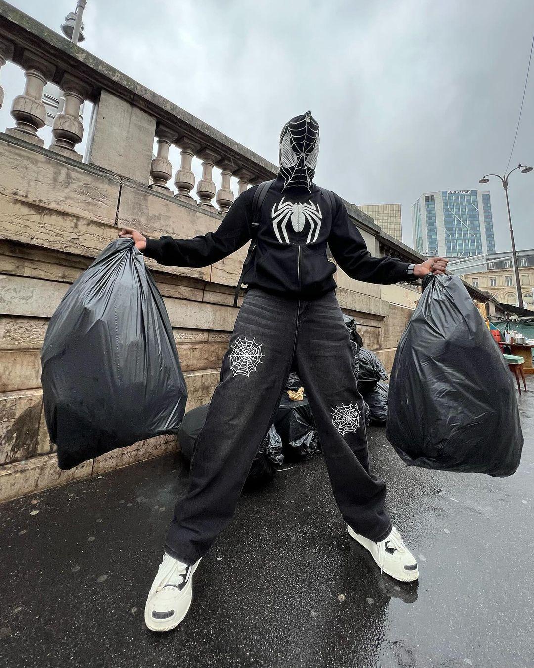 A person wearing a Spider Web Embroidery Eye Viewable Hoodie by TALISHKO, holding trash bags.
