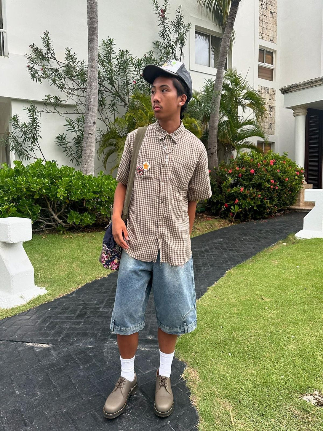 Model wearing a TALISHKO short-sleeved shirt with denim shorts and a cap in a casual setting.