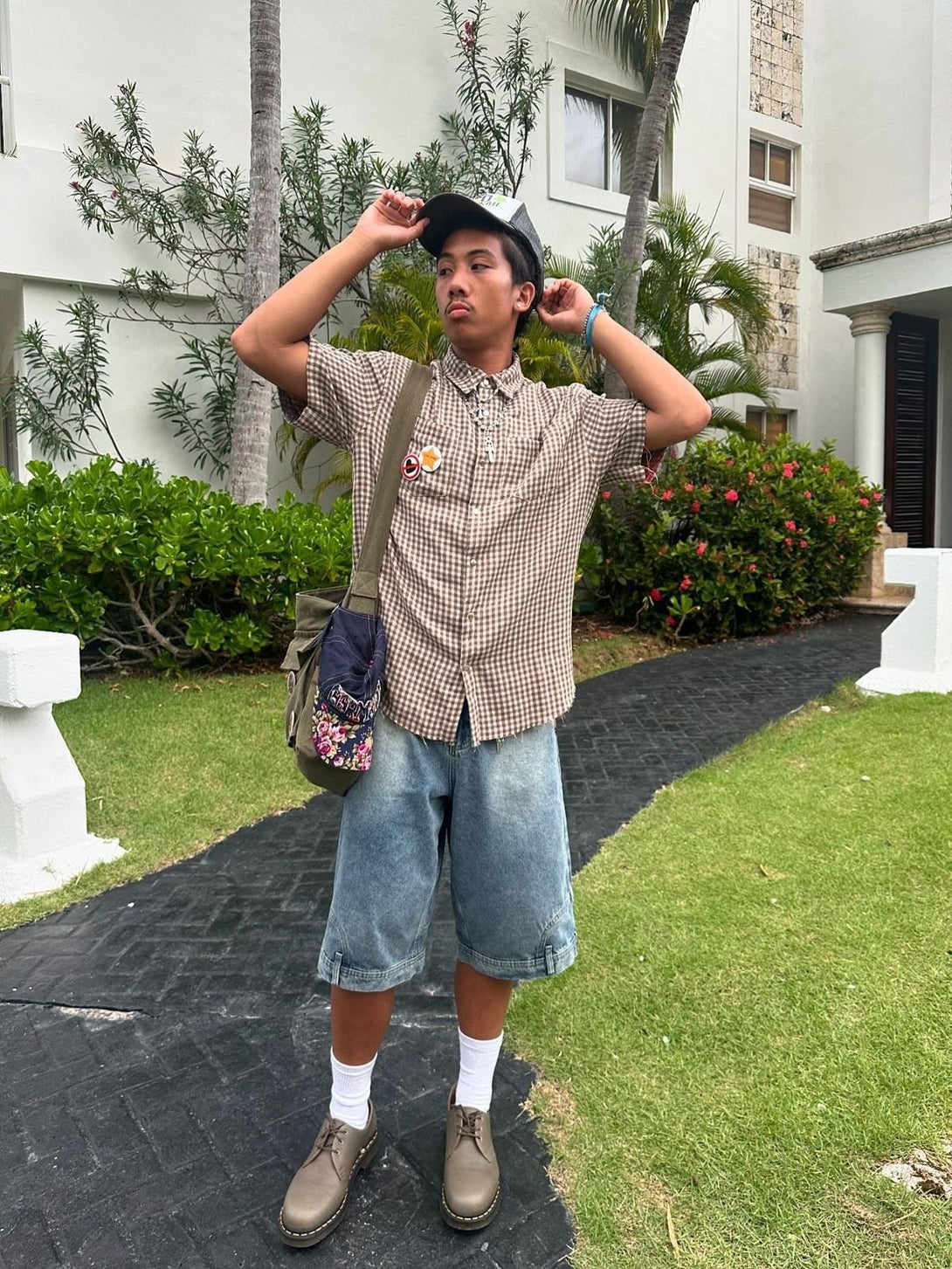 Model in a checkered TALISHKO shirt and denim shorts, adjusting a cap in a garden setting.