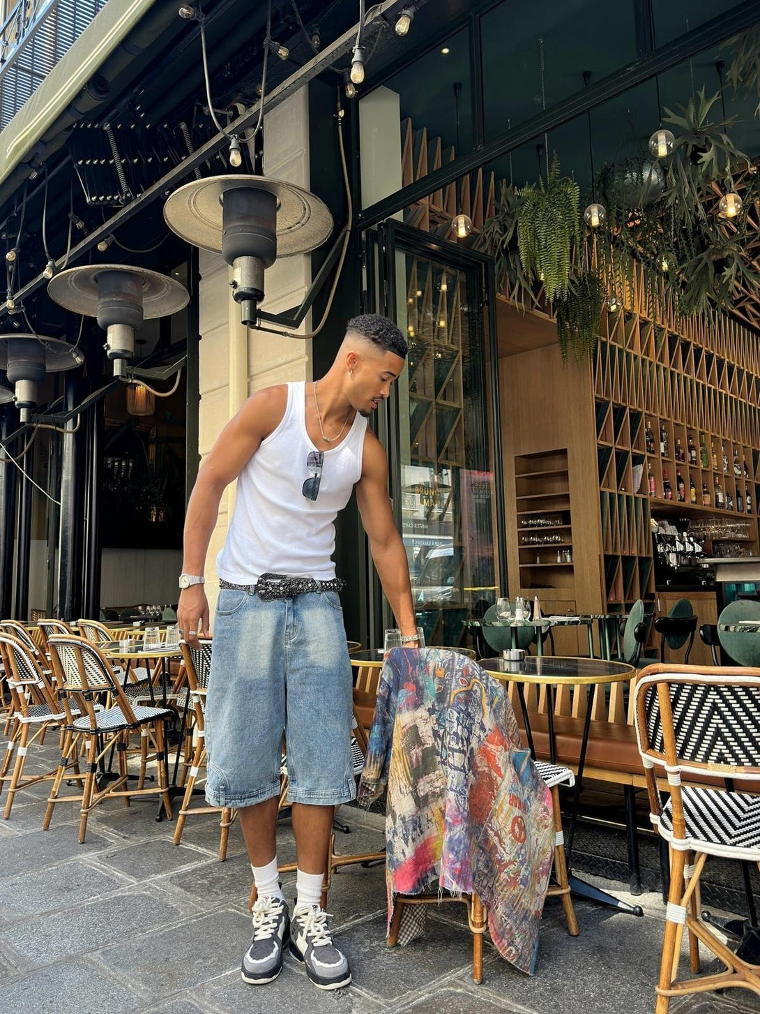 Model in a vibrant TALISHKO short-sleeved shirt and denim shorts, standing outside a café.