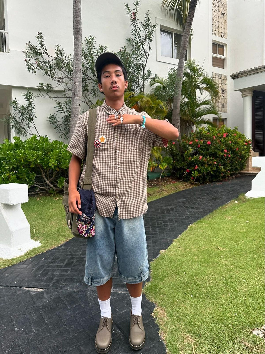 Model sporting a TALISHKO checkered short-sleeved shirt with denim shorts in a garden setting.