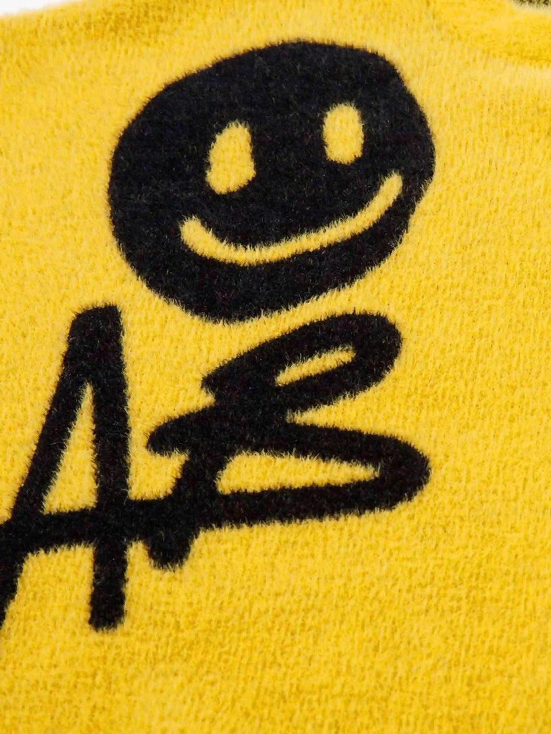Zoomed-in texture of a yellow TALISHKO sweater featuring a playful black smiley face design.