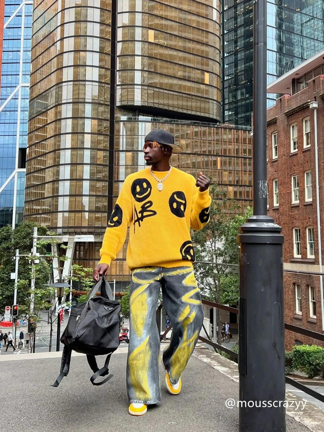 A model in a vibrant TALISHKO sweater with smiley graphics, paired with artistic jeans on a city street.