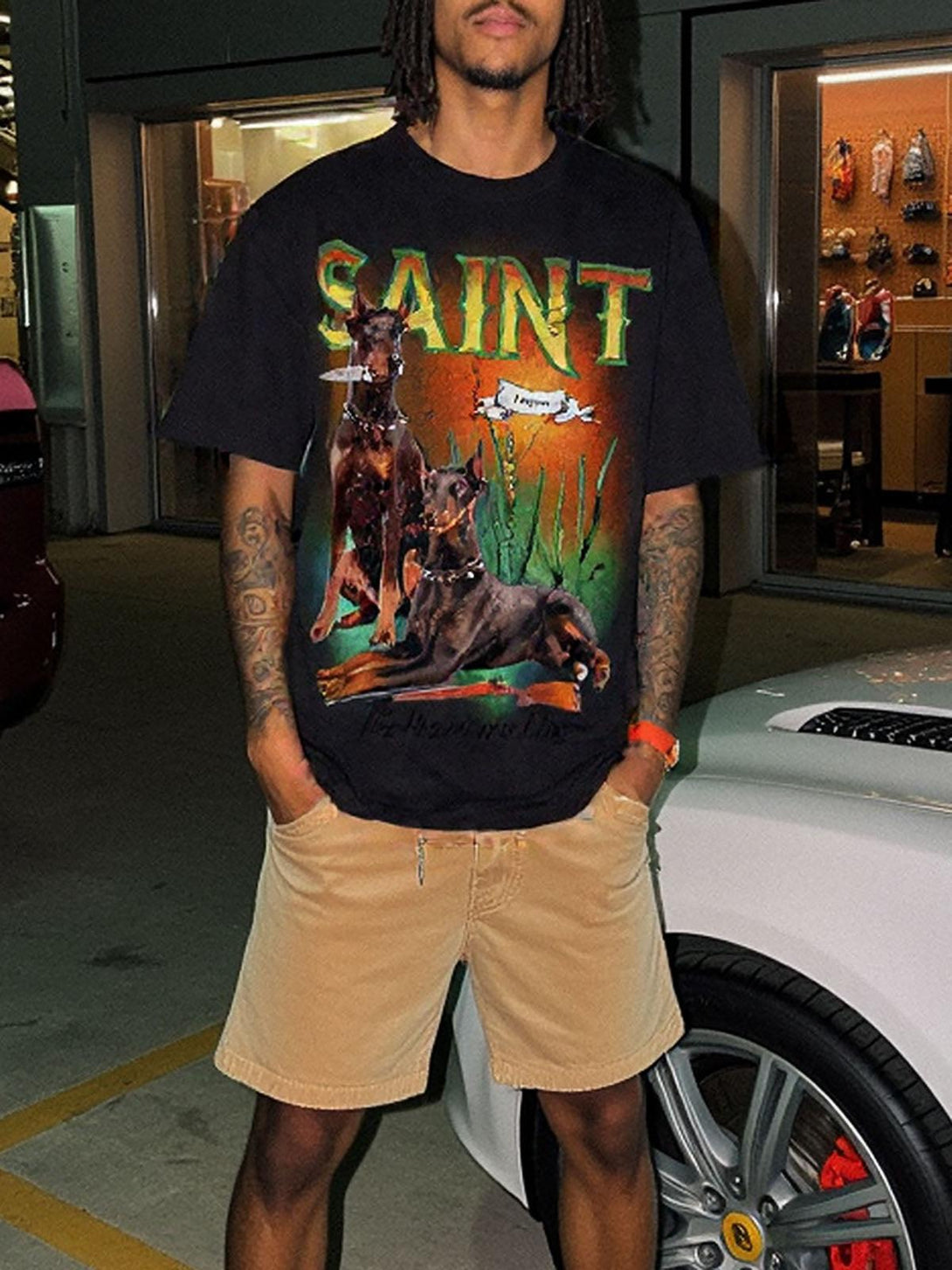 Model posing in a TALISHKO vintage Doberman print T-shirt and beige shorts near a car.