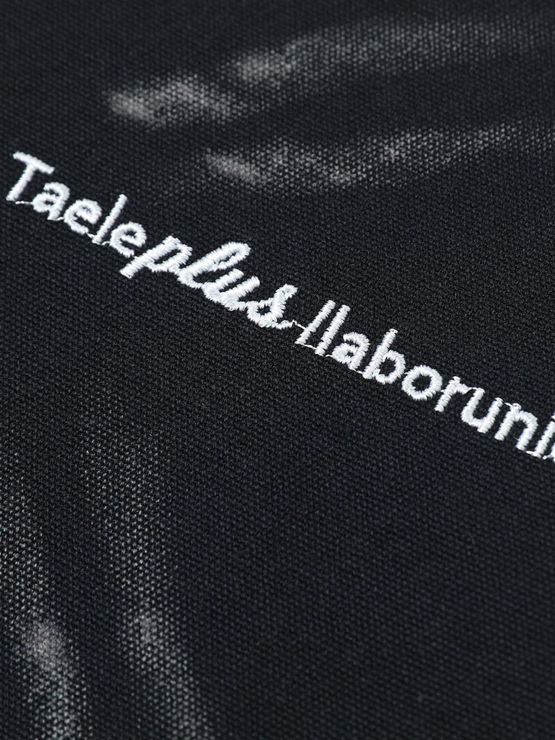 Intricate look at TALISHKO's black shirt fabric texture with white embroidered text detail.